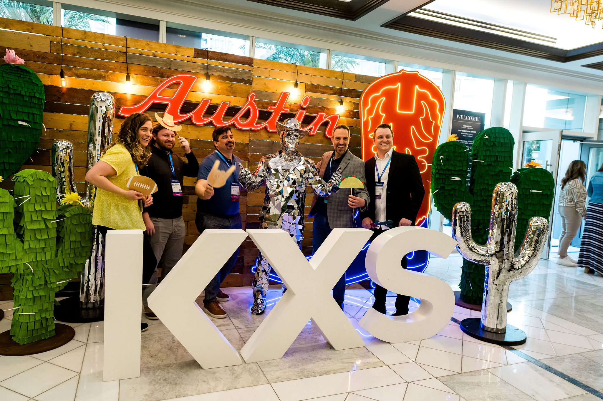 Kinexions A group of Kinexions attendees pose with a mirrored cowboy performer and props in front of a neon-lit “Austin” sign and giant “KXS” letters at a themed photo station at one of many corporate events at the Fairmont Austin.