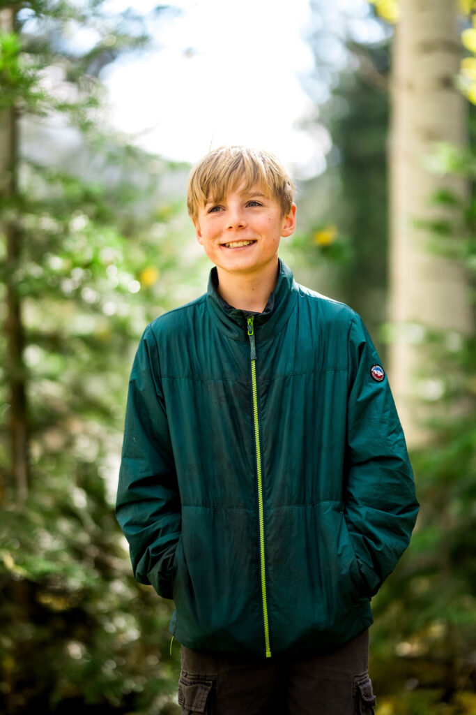 best-locations-for-family-portraits-in-south-lake-tahoe-60 A boy in a green jacket smiles in a sunlit forest near the Christmas Valley trailhead.