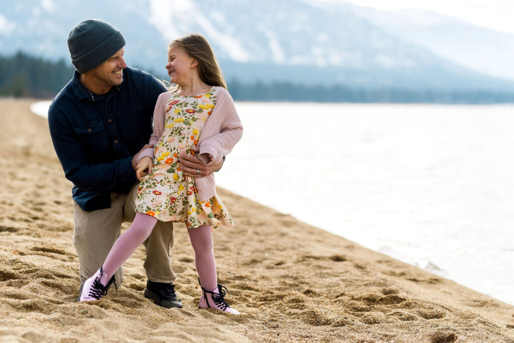 best-locations-for-family-portraits-in-south-lake-tahoe-68 A father kneels on the beach at Nevada Beach in Stateline, Nevada, smiling at his daughter near the water.