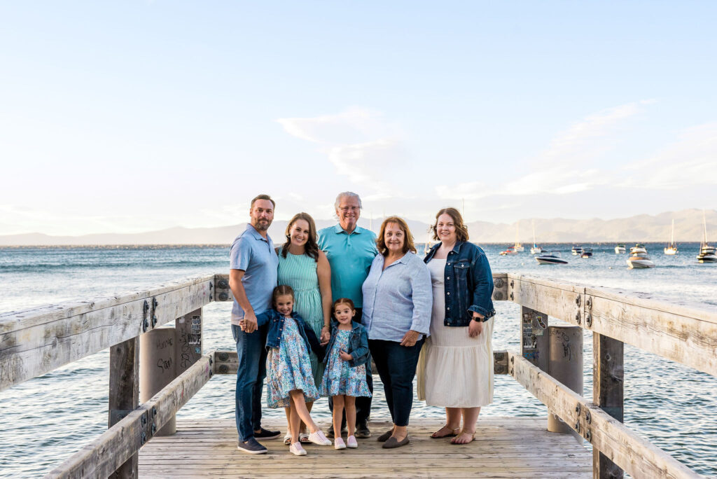 best-locations-for-family-portraits-in-south-lake-tahoe-95 A multigenerational family of seven stands on the wooden dock at Valhalla in South Lake Tahoe, with calm water, boats, and mountains behind them.
